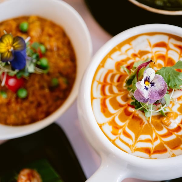 Two bowls of Indian curry, one with a decorative cream swirl and edible flower, the other with peas.