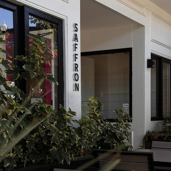 Exterior of Saffron Indian restaurant with its name vertically on a white pillar, surrounded by green plants.