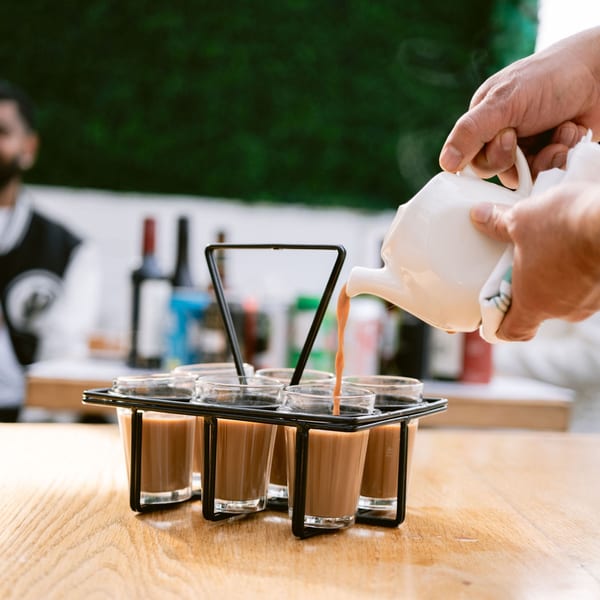 A hand pouring Saffron's Indian chai from a white teapot into small glasses in a metal carrier.