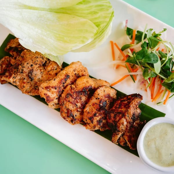 Grilled chicken pieces, lettuce wraps, a side salad, and a green dipping sauce on a white plate.