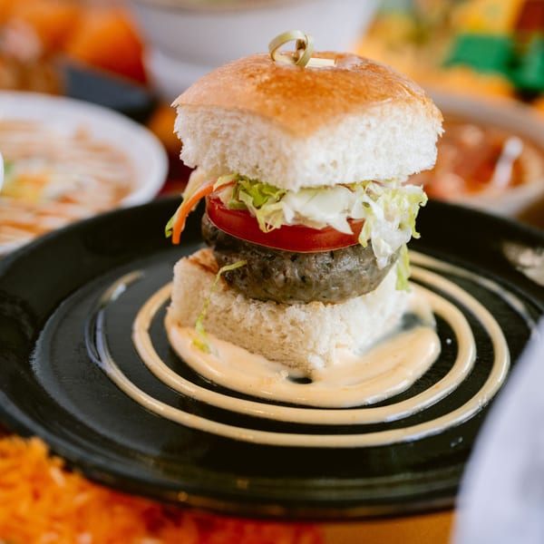 A Saffron Indian slider with a patty, tomato, lettuce, and sauce on a black plate.
