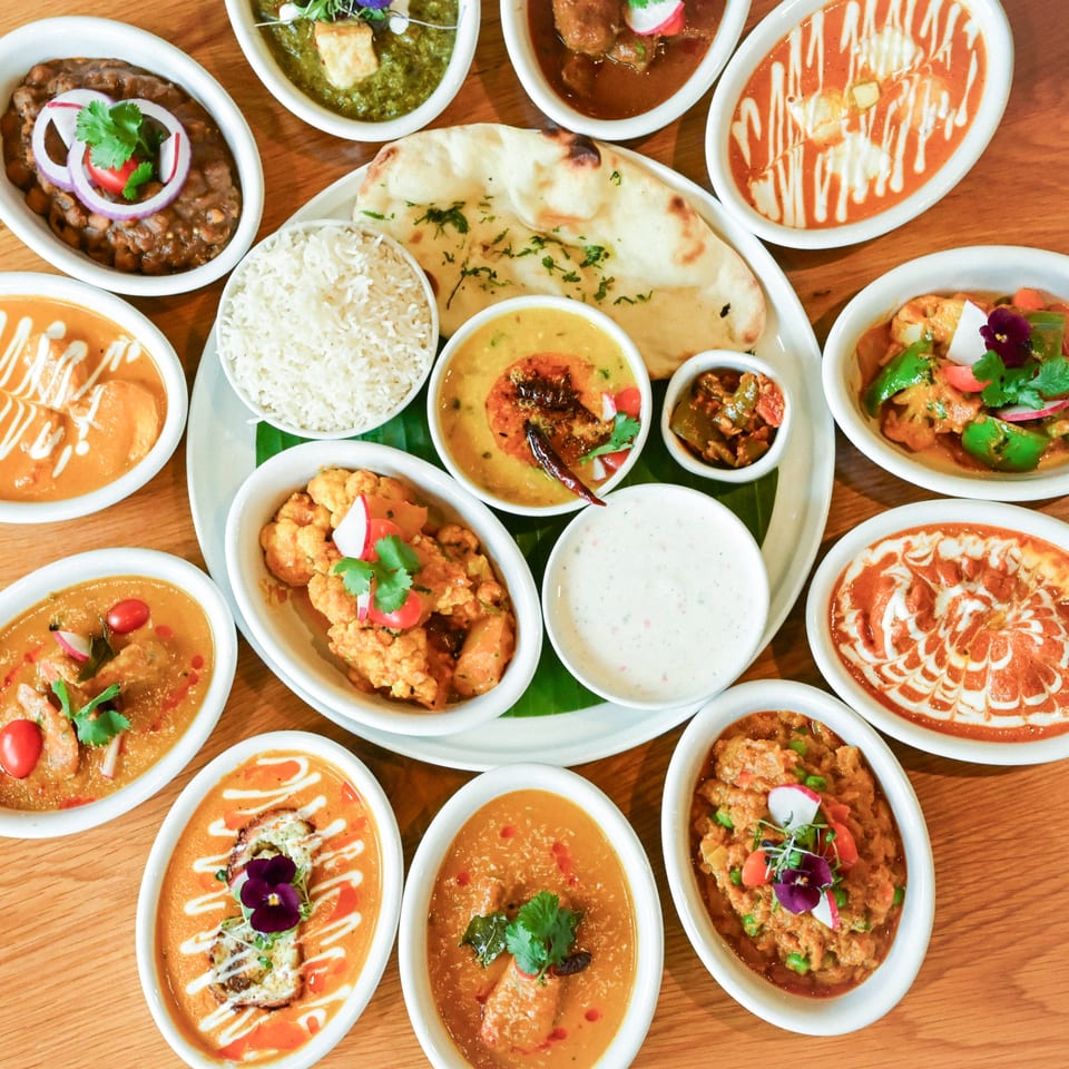 A vibrant spread of Indian dishes, including curries, rice, naan, and raita, on a wooden table.