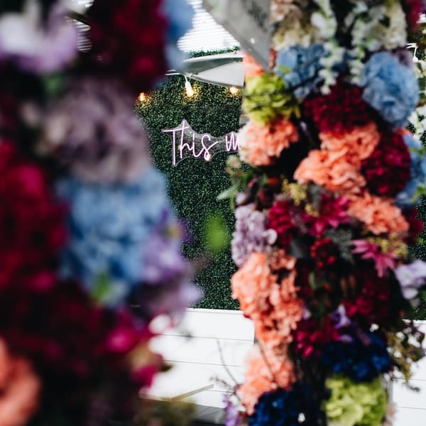 A neon sign reading "This Way" on a green hedge wall, framed by colorful floral arrangements.