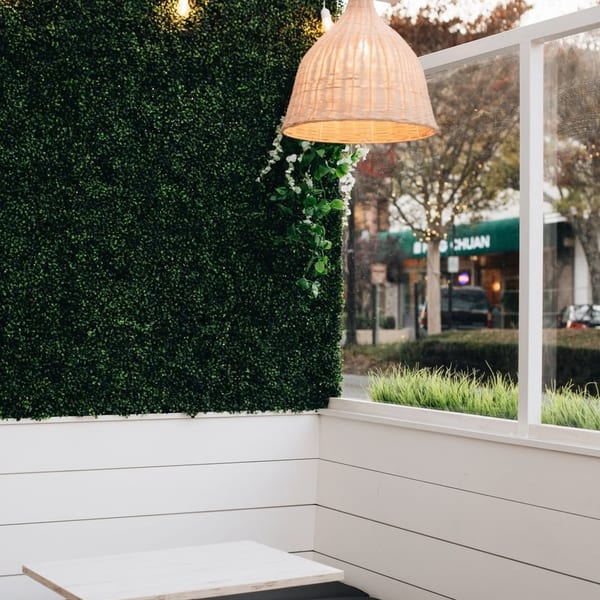 Outdoor dining area with a green hedge wall, white paneling, and a woven light fixture.