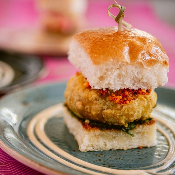 A Saffron Indian vada pav, a fried potato patty in a bun, on a blue plate.
